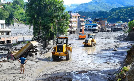 Equipamentos pesados ​​de construção, incluindo carregadeiras e um caminhão basculante, operam em um assentamento coberto de lama em Nuwakot, Nepal, com prédios danificados e montanhas ao fundo após enchentes repentinas.
