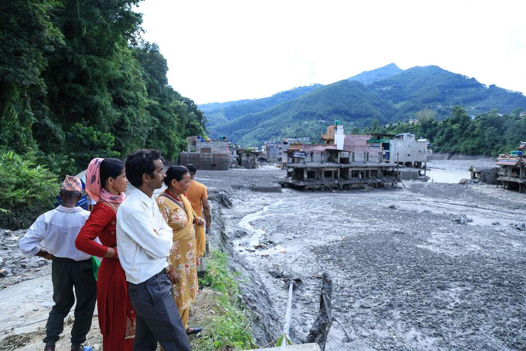 Des villageois népalais se tiennent au bord d'une rive boueuse dans le district de Nuwakot, observant les bâtiments endommagés par les inondations et le sol recouvert de boue après les crues soudaines du 26 août 2026.