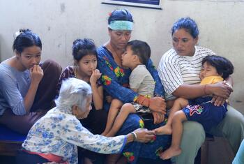 A Nepali family of six, including an elderly woman and several young children, sits together on a bench inside a school building in Nuwakot District, Nepal, after floods displaced them from their home.
