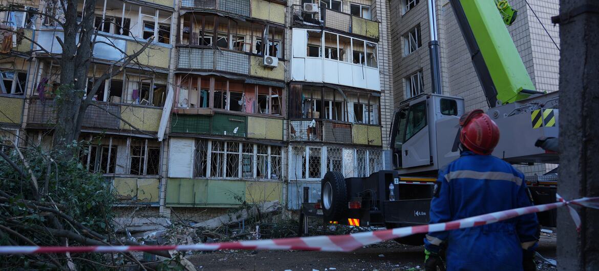 A rescue worker in a red helmet and blue overalls stands before a severely damaged apartment building in Kyiv’s Solomianskyi district, with a green crane positioned beside the structure amid debris and shattered windows.