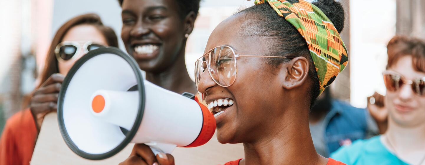Une femme noire souriante, vêtue d'un t-shirt orange et d'un foulard, parle dans un mégaphone blanc lors d'une manifestation pacifique dans le centre-ville de Kansas City, entourée de sympathisants brandissant des pancartes.