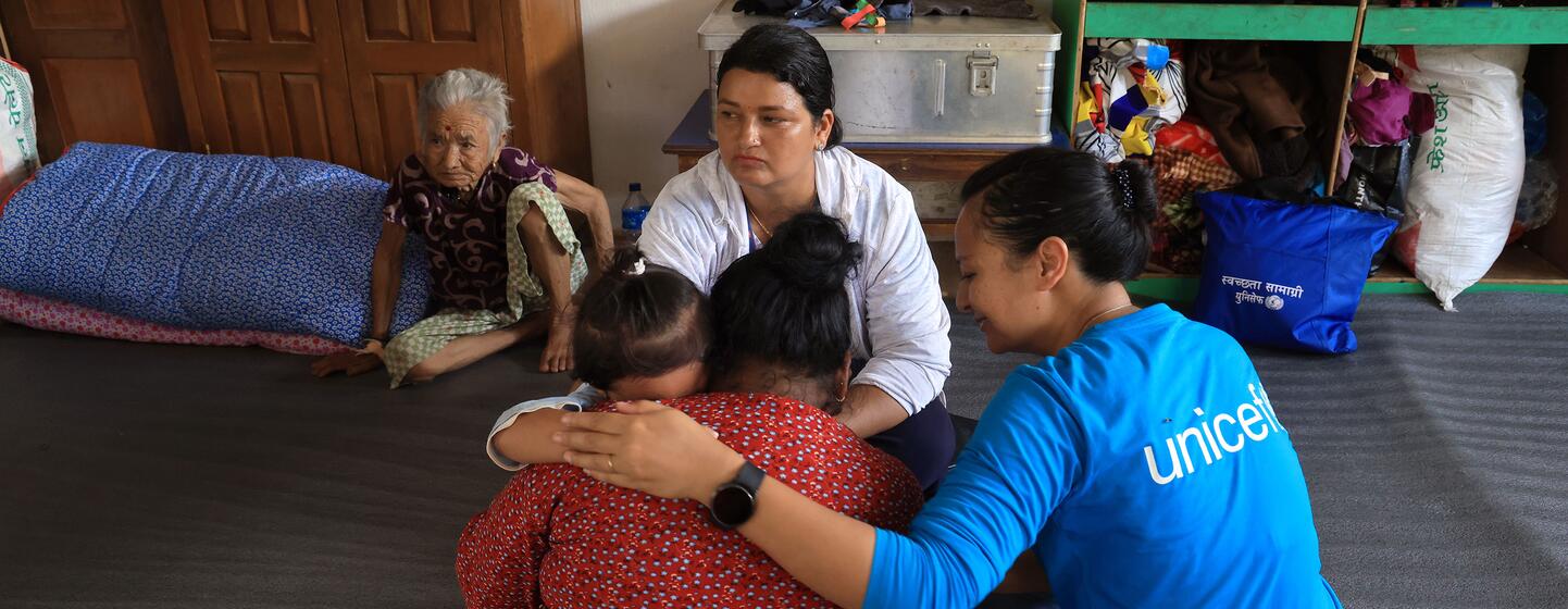 À Nuwakot, au Népal, une employée de UNICEF vêtue d'une chemise bleue réconforte une femme en détresse qui tient deux enfants dans ses bras, allongée sur le sol d'un refuge, sous le regard d'une femme âgée et d'un autre adulte.