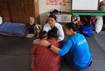 A UNICEF worker in a blue shirt comforts a distressed woman holding two children on the floor of a shelter in Nuwakot, Nepal, while an elderly woman and another adult look on.