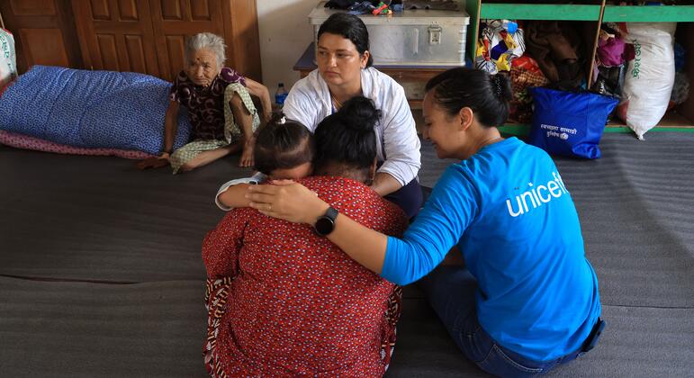 À Nuwakot, au Népal, une employée de UNICEF vêtue d'une chemise bleue réconforte une femme en détresse qui tient deux enfants dans ses bras, allongée sur le sol d'un refuge, sous le regard d'une femme âgée et d'un autre adulte.