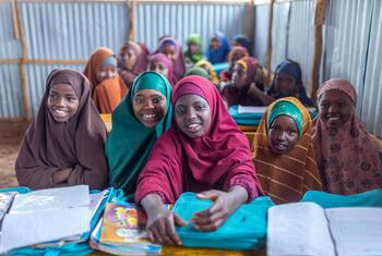 Smiling Somali girls in colorful headscarves sit at desks with books and blue folders inside a corrugated-metal UNICEF-supported Waaberi IDP school classroom in Baidoa.