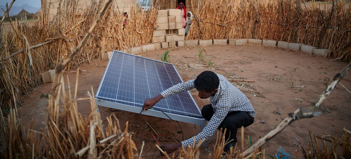 A young African man crouches to install a solar panel in a dry yard beside a block building and thatched shelter in Kassala, Eastern Sudan.