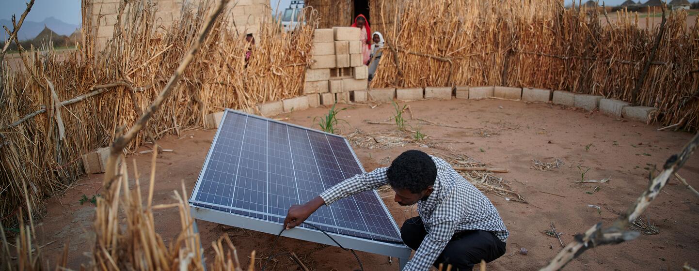 Un joven africano se agacha para instalar un panel solar en un patio seco junto a un edificio de bloques y un refugio con techo de paja en Kassala, Sudán Oriental.