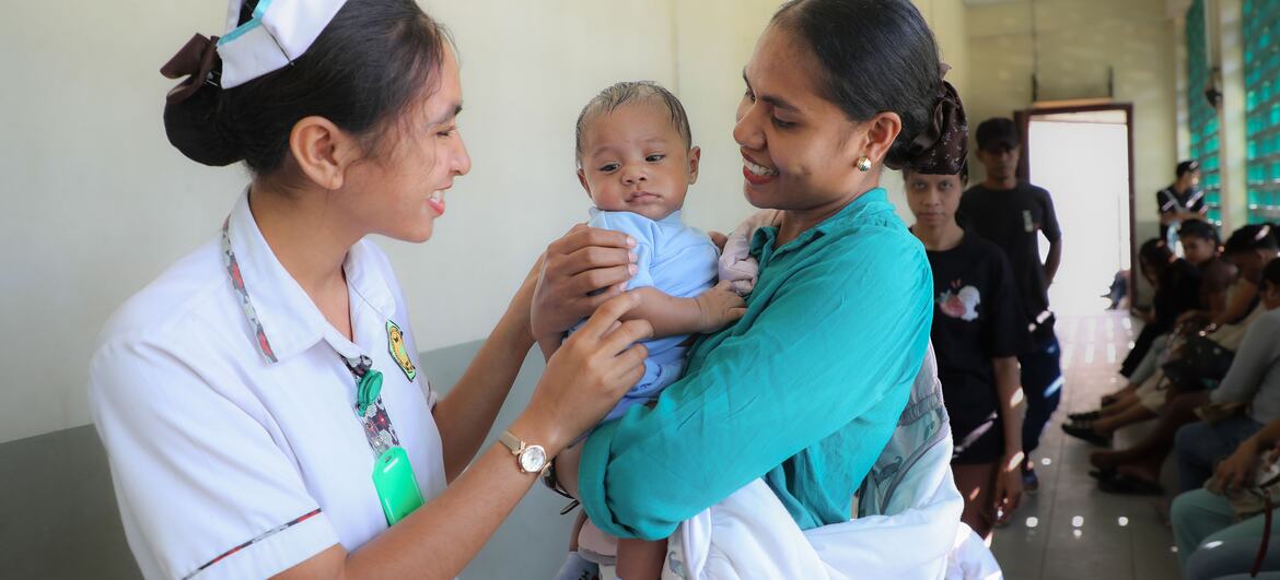 A nurse in a white uniform and cap hands a smiling 3-month-old baby to his mother at the Formoza Community Health Centre in Timor-Leste, with other people waiting along the corridor behind them.