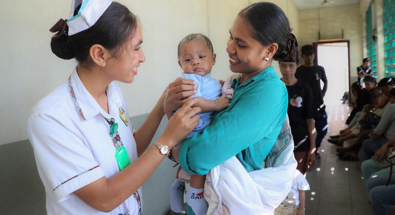 Una enfermera con uniforme y gorro blancos entrega a su madre a un bebé sonriente de 3 meses en el Centro de Salud Comunitario de Formoza, en Timor Oriental, mientras otras personas esperan en el pasillo detrás de ellos.