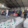 Personal uniformado y de civil manipula bidones de plástico blanco y cajas de cartón dentro de un gran almacén en el Área de Concentración Humanitaria de Katmandú durante el transporte de suministros para refugios de emergencia facilitado por el PMA.