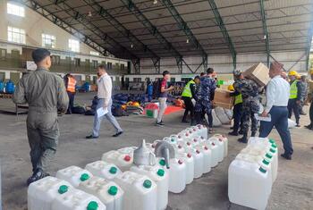 Personnel in uniform and civilian attire handle white plastic jerrycans and cardboard boxes inside a large warehouse at the Kathmandu Humanitarian Staging Area during WFP-facilitated emergency shelter supply transport.