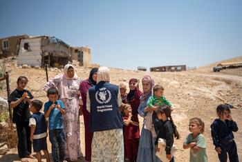 Un miembro del personal del PMA , que lleva un chaleco azul con el logotipo del Programa Mundial de Alimentos, se dirige a un grupo de mujeres y niños palestinos en Sada Al Tha'la, en la zona C de Cisjordania, cerca de refugios improvisados.