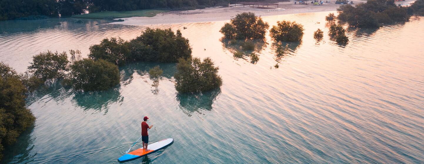 Vista aérea de una persona con una camisa roja de pie sobre una tabla de paddle surf naranja y azul, deslizándose por las tranquilas aguas turquesas entre islas de manglares al atardecer, con una playa y una costa boscosa visibles a lo lejos.