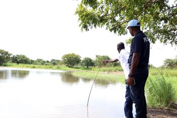 A man in a light blue cap and dark shirt stands at the edge of a newly built artificial lake in Terekeka, South Sudan, while another man points toward the water; palm trees and green grass line the far bank under a bright sky.