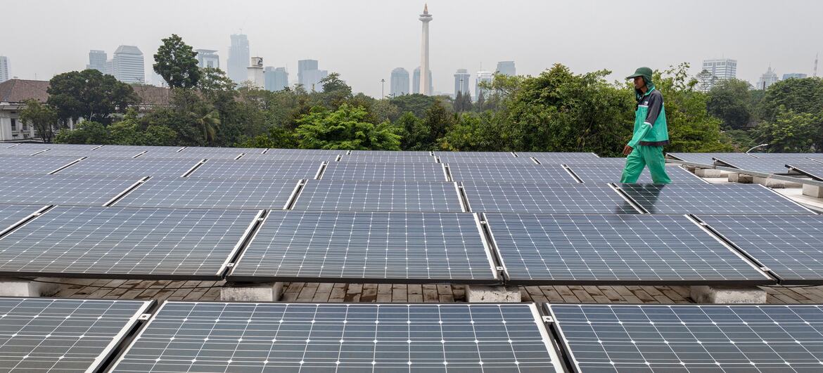 A worker in green uniform and cap walks across rows of rooftop solar panels at the Istiqlal Mosque in Central Jakarta, with the hazy Jakarta skyline and National Monument visible in the background.