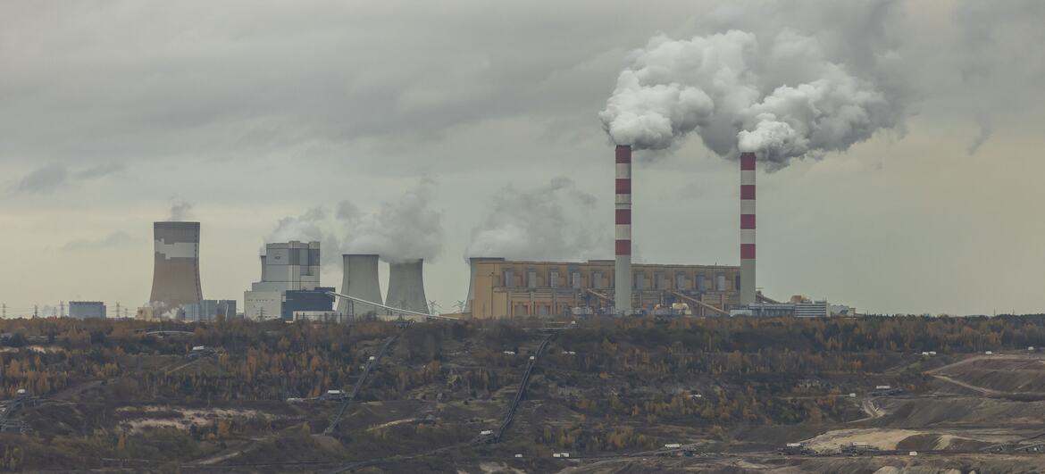 Wide view of the Belchatow coal-fired power station in Zlobnica, Poland, showing its cooling towers and tall striped chimneys releasing plumes of smoke over a sprawling open-pit mining landscape under an overcast sky.