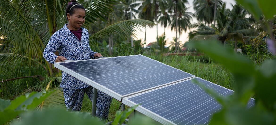 Una mujer camboyana con una blusa estampada de flores se encuentra junto a dos paneles solares montados sobre una estructura metálica en una zona rural rodeada de palmeras de coco y exuberante vegetación.