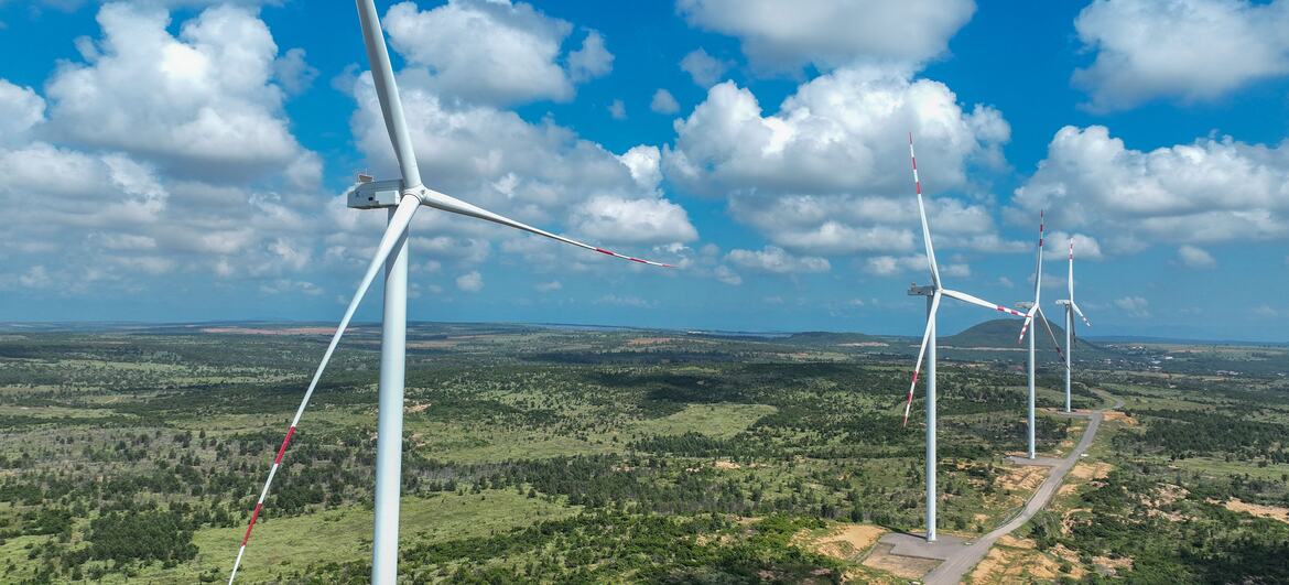 Wind turbines with red and white blades stand in a line across a green rural landscape under a bright blue sky with scattered clouds.