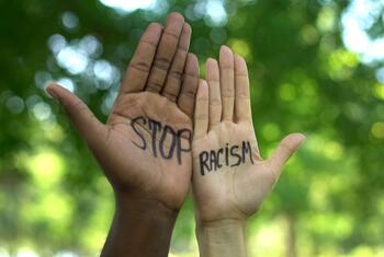 Duas mãos estendidas, uma de pele escura e outra de pele clara, com as palavras "PARE" e "RACISMO" escritas com marcador preto nas palmas, sobre um fundo desfocado de folhagem verde.
