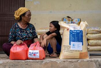 An elderly Nepali woman wearing a yellow headscarf and a young girl sit on a low stone ledge beside red plastic bags labeled Australian Aid and large sacks of rice during WFP-supported food distribution in Archale, Nuwakot District, Nepal.