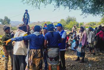 Un travailleur humanitaire de l'OIM, coiffé d'une casquette bleue et vêtu d'un gilet, s'entretient avec un groupe d'hommes et de garçons rassemblés sur un terrain rocailleux en extérieur au Yémen, lors d'une activité d'assistance humanitaire.