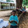Un jeune garçon vêtu d'un sweat-shirt turquoise lit un livre ouvert, assis près des racines d'un arbre. Derrière lui, d'autres enfants étudient à l'ombre d'un grand arbre, à côté d'une école endommagée et sans toit, à Nacala, au Mozambique.