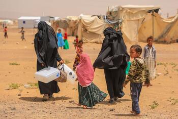 Deux femmes vêtues de burqas noires intégrales transportent des bidons d'eau en plastique blanc tout en marchant pieds nus avec plusieurs enfants à travers un campement sablonneux parsemé de tentes en toile.