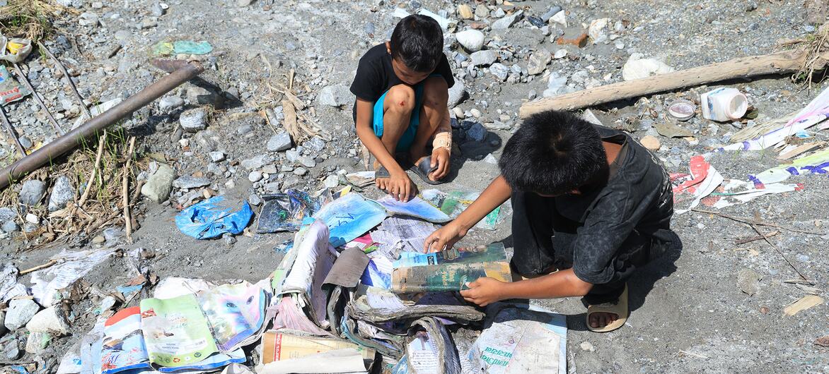 Two children search through scattered papers and debris on rocky, flood-affected ground. The surroundings show signs of flood damage with washed-up material and rubble.