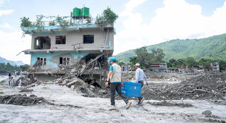 Two men, one wearing a light vest and blue cap, carry a blue plastic container across a muddy debris field beside a damaged two-story house with an eroded foundation in a hilly area.