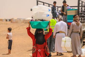 IOM staff in high-visibility vests assist as a woman in a red dress and black headscarf carries stacked water jerrycans on her head while families receive NFI and shelter supplies from a truck at a displacement site in Yemen.