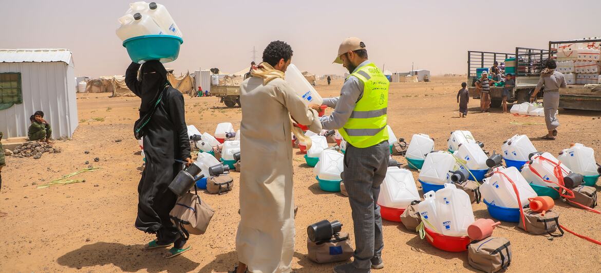 IOM aid worker in high-visibility vest hands a water jerrycan to a man in a light robe at a displacement camp in Yemen, while a woman in black carries stacked water containers on her head; numerous jerrycans and backpacks are arranged on the sandy ground with tents and a truck in the background.