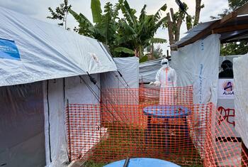 Un agent de santé en tenue de protection complète se tient derrière une barrière de sécurité orange sur un site d'extension d'un centre de traitement Ebola soutenu par l'OMS à Lita, avec des tentes portant les marques UNICEF et ALIMA visibles.