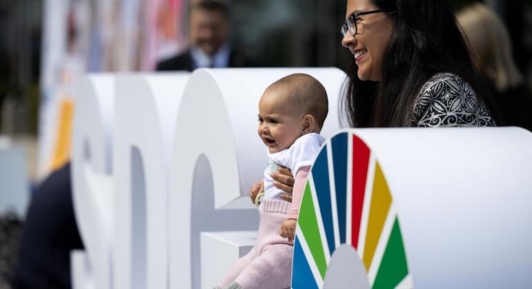 Priscila Franco holds her 5-month-old daughter Priscilla, seated on large white SDG letters beside a colorful UN sustainable development goals emblem at UN Headquarters.