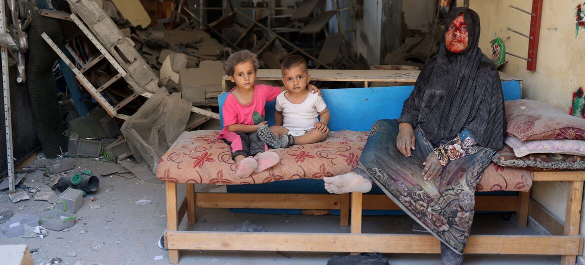 A wounded woman wearing a black headscarf sits barefoot on a mattress with two young children beside her in a damaged room filled with rubble and debris in Burij, Gaza Strip.