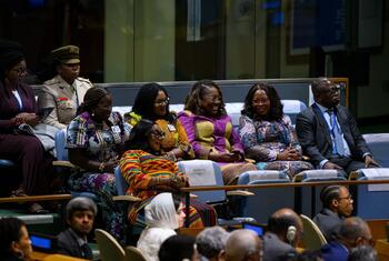 Delegates, including women in colorful African-print dresses and a uniformed officer, are seated in a gallery area at the United Nations during an official session.