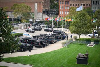 Elevated view of the United Nations Headquarters plaza in New York with rows of black official SUVs, national flags, security personnel near vehicles, and a bronze sculpture on a green lawn.