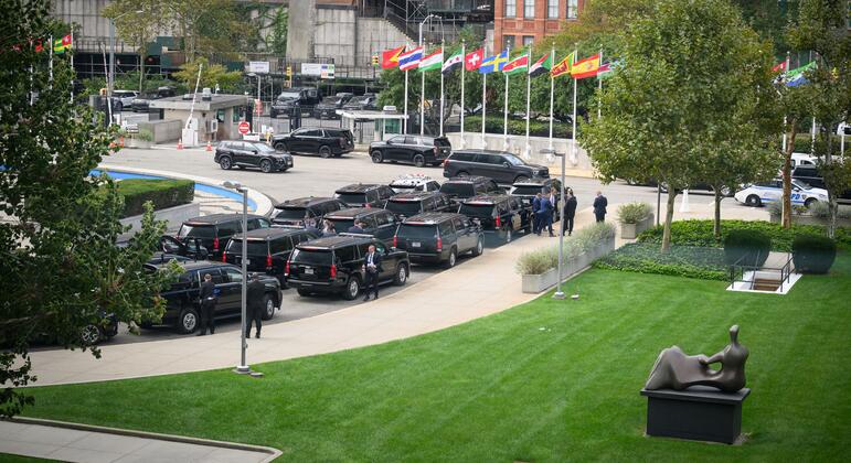 Elevated view of the United Nations Headquarters plaza in New York with rows of black official SUVs, national flags, security personnel near vehicles, and a bronze sculpture on a green lawn.