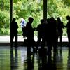 Silhouetted delegates and staff stand in a reflective, glass-walled UN Headquarters lobby as green trees are visible outside on the first day of the 81st General Assembly debate.