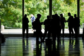 Silhouetted delegates and staff stand in a reflective, glass-walled UN Headquarters lobby as green trees are visible outside on the first day of the 81st General Assembly debate.