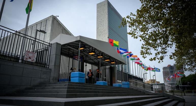 Штаб-квартира United Nations в Нью-Йорке с рядами национальных флагов, гранитными ступенями, навесом у входа и несколькими сотрудниками у ворот пасмурным утром.