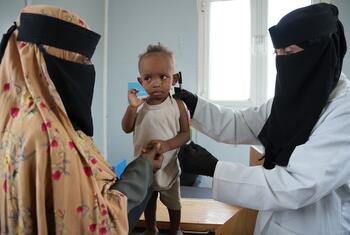 A veiled female health worker in a white coat measures the arm of a thin Yemeni boy with a MUAC tape while his veiled mother holds him at a clinic in Aden.