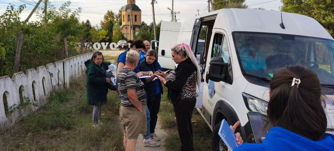 UN OCHA field workers in blue shirts speak with Ukrainian women and men beside a white aid van on a rural roadside, holding documents and a tablet, with a church tower and fence in the background.