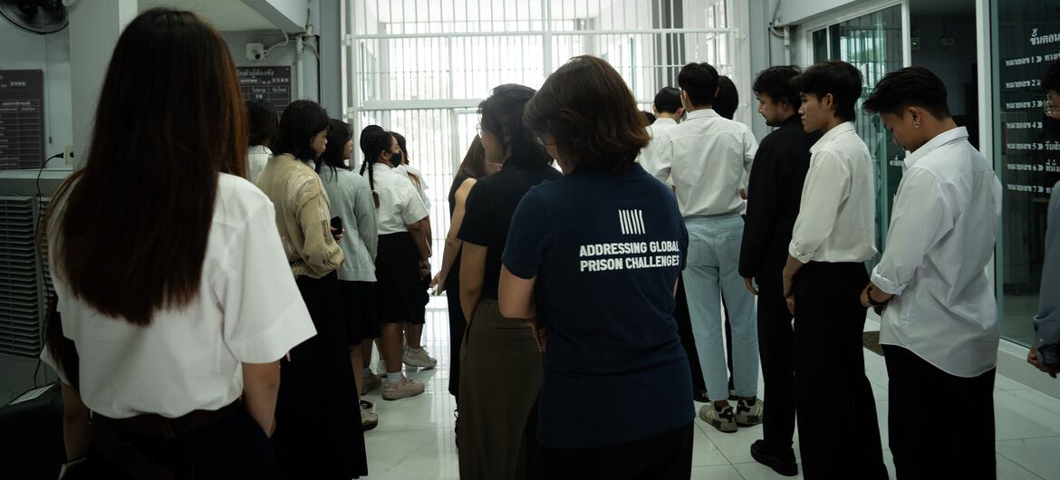 Young Asian university students stand in a line inside a correctional facility visiting room, near a barred gate and a wall sign reading SEARCHING ROOM; one female student wears a dark blue shirt with white lettering.