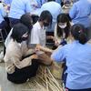 Young Asian female university students wearing masks sit on the floor with women in light blue uniforms, handling bamboo strips and weaving baskets in a craft workshop.