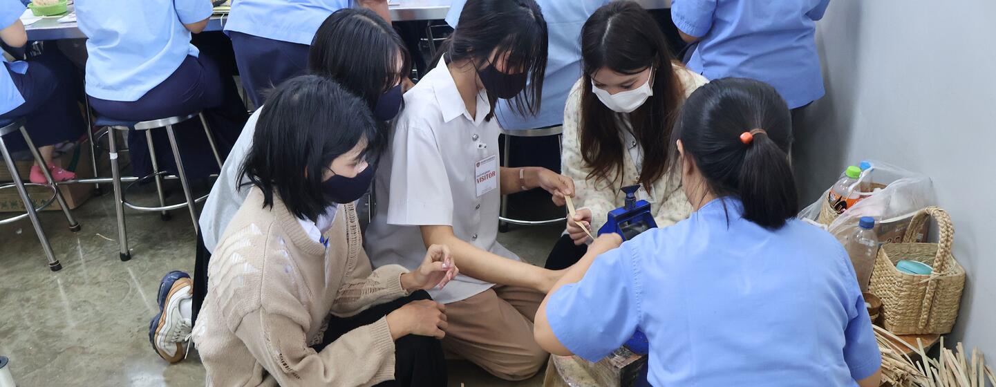 Young Asian female university students wearing masks sit on the floor with women in light blue uniforms, handling bamboo strips and weaving baskets in a craft workshop.
