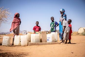 African women and children stand at an outdoor water point with white jerrycans, including one marked IOM, in a dry camp setting with tents and bare trees.