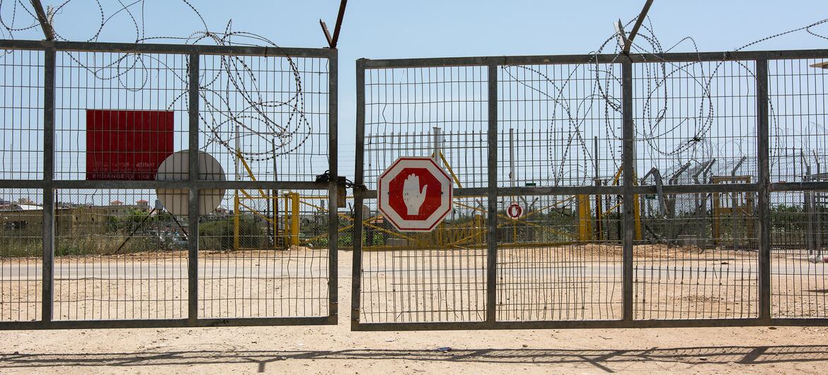 Una puerta metálica cerrada con alambre de púas en un puesto de control fronterizo israelí-palestino bloquea un camino de tierra, y una señal roja de stop es visible detrás de la valla bajo un cielo despejado.