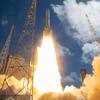 A launch vehicle lifts off from a launch pad with bright engine exhaust and billowing smoke under a partly cloudy blue sky.