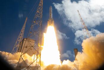 A launch vehicle lifts off from a launch pad with bright engine exhaust and billowing smoke under a partly cloudy blue sky.