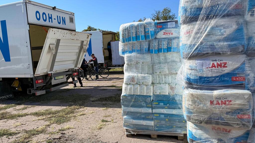 Un camion des ONU OHH-UN, porte arrière ouverte, décharge des palettes de produits d'hygiène et de couches KANZ sur un site de distribution en plein air dans la région de Kherson ; des travailleurs et des vélos se tiennent à proximité sur un sol en terre battue sous un ciel bleu clair.